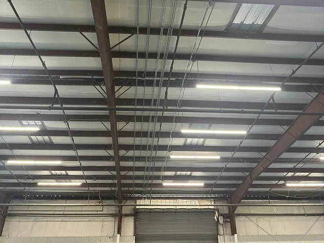 Linear LED fixtures and conduit runs along the structural steel of a warehouse ceiling